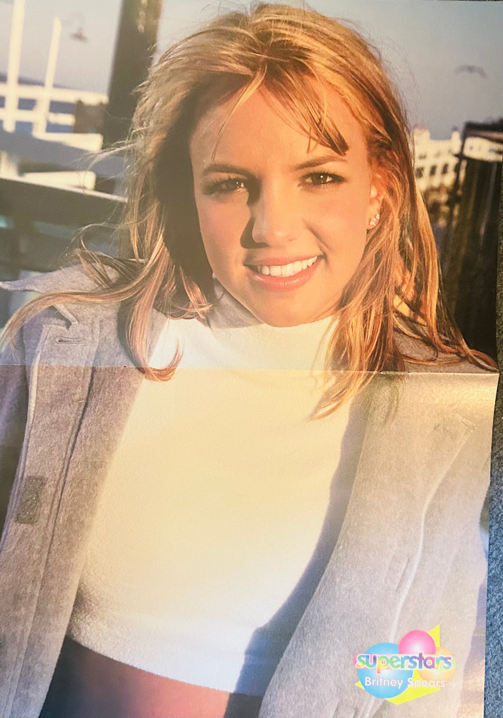 Superstars magazine double-sided poster side A featuring Britney Spears outdoor candid portrait in white turtleneck and grey blazer with wind-swept hair smiling against bright sky and urban backdrop, Superstars Britney Spears logo bottom right