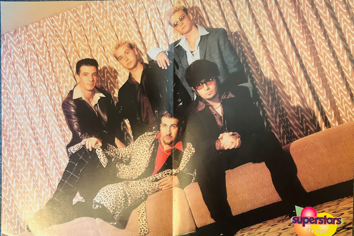 Superstars magazine double-sided poster side B featuring all five NSYNC members in bold early 2000s fashion including leather jackets leopard print sunglasses and plaid trousers posed in warmly lit hotel lounge with chevron curtains, Superstars N Sync logo bottom right