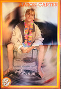 Pop Star! magazine double-sided poster side A featuring Aaron Carter barefoot sitting on metal chair wearing khaki jacket and colorful tropical flower lei with spinning vinyl records in background, AARON CARTER in bold orange font top center, orange border, Pop Star logo bottom left