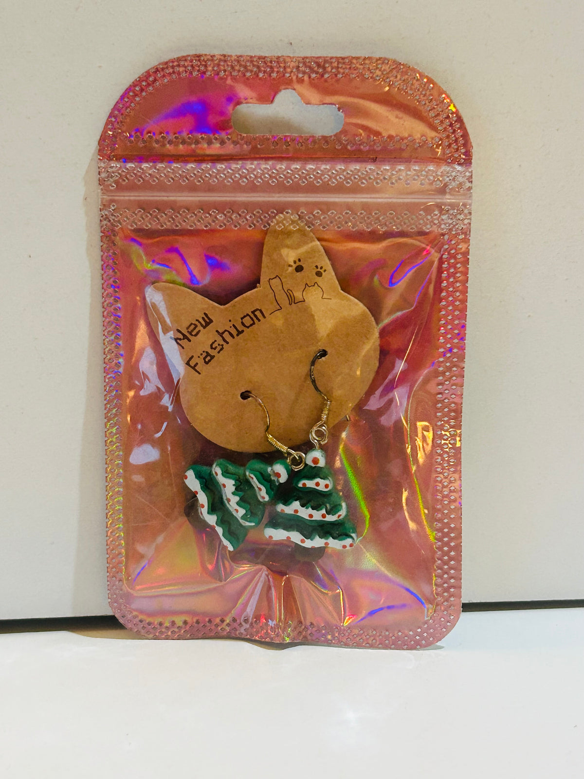 Pair of green Christmas tree clay charm drop earrings with white trim and red ornament dots on gold fish hook wires, sealed in holographic pink zip bag with New Fashion cat display card