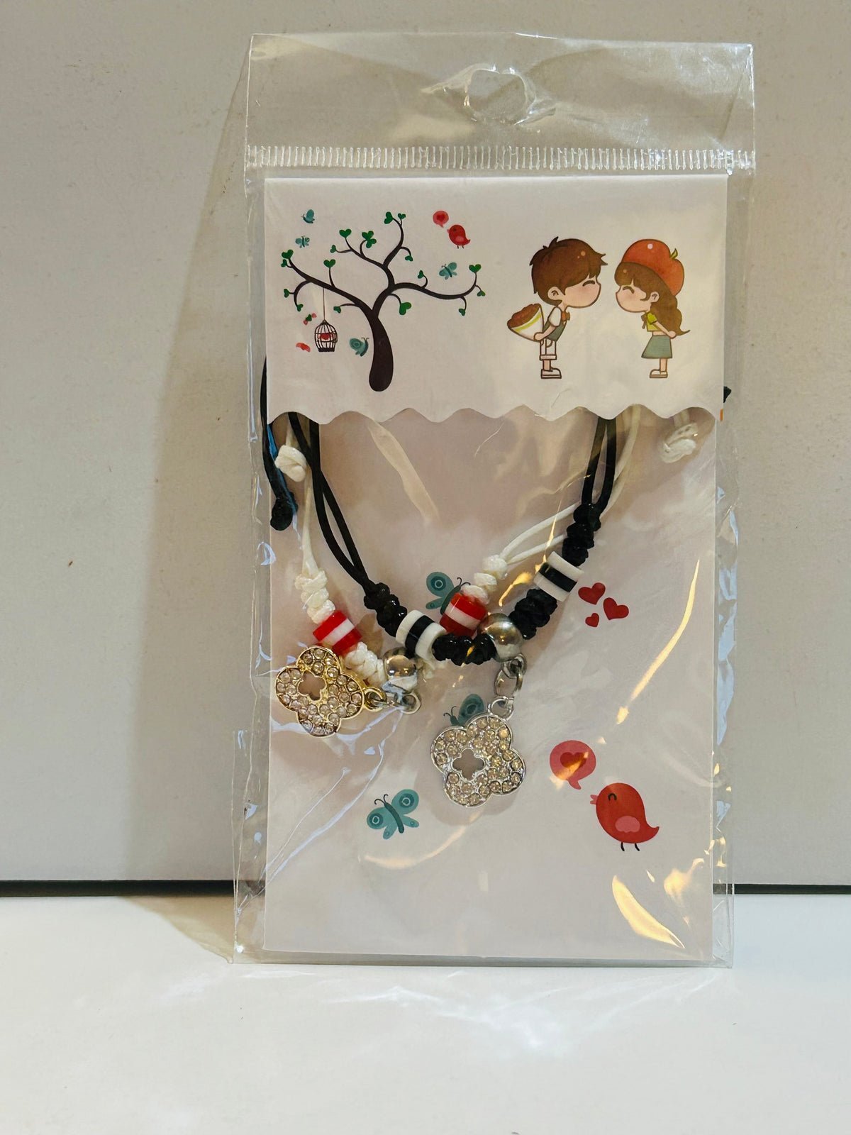 Two matching black braided cord bracelets with red, white, and black beads and rhinestone flower charms, sealed in packaging with illustrated couple display card