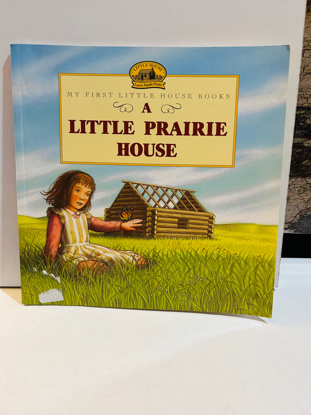 A Little Prairie House 9x9 paperback cover from My First Little House Books series by Laura Ingalls Wilder, featuring young Laura in striped dress sitting in golden prairie grass with monarch butterfly, log cabin being built in background, 1998 HarperCollins