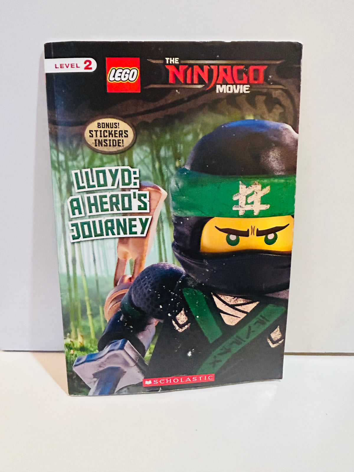 The LEGO Ninjago Movie Lloyd A Hero's Journey 2017 Scholastic Level 2 reader 6x9 paperback cover featuring Lloyd the Green Ninja in black and green armor with yellow face mask in bamboo forest, Bonus Stickers Inside badge, red LEGO logo