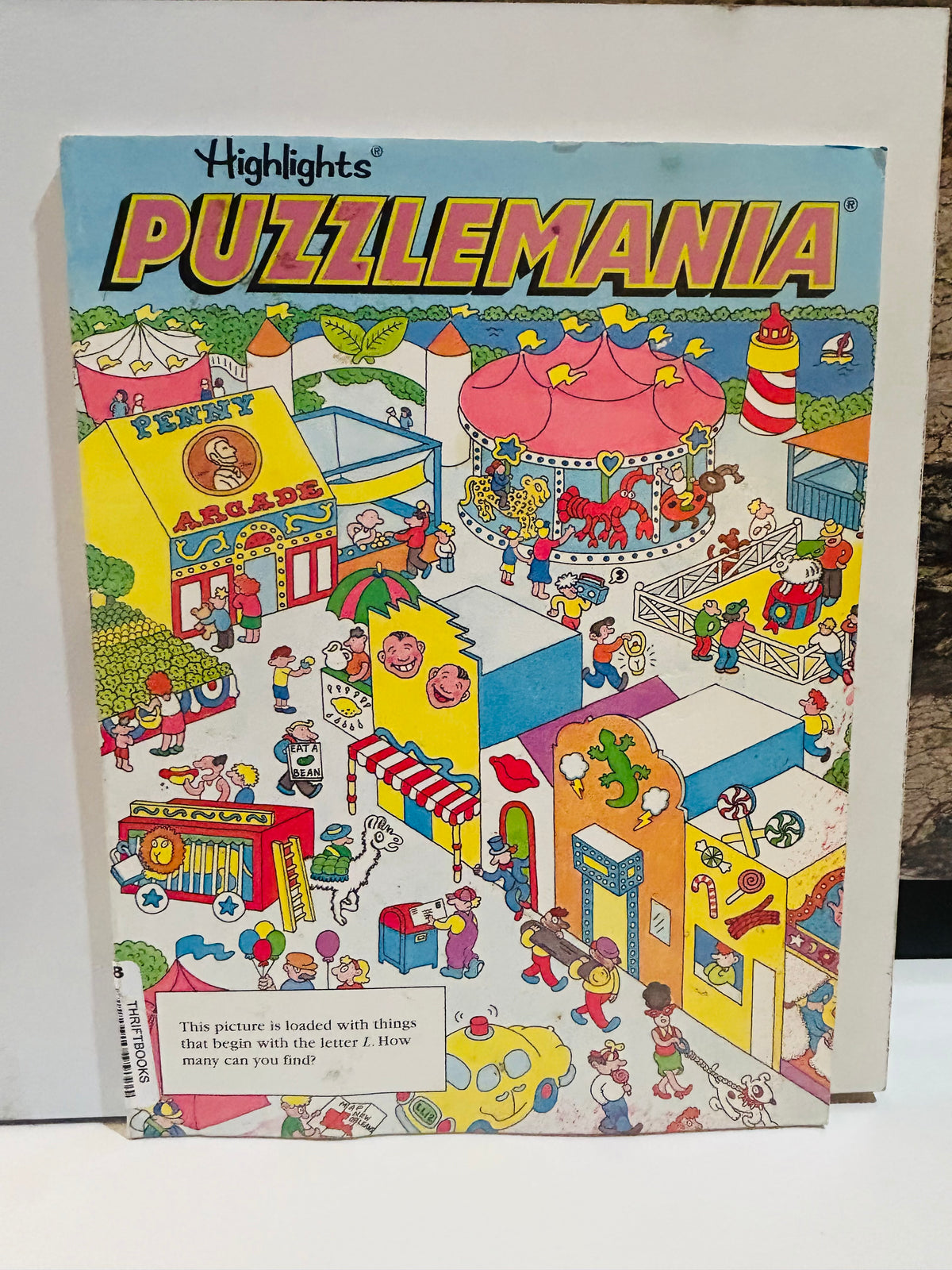 Highlights Puzzlemania Book 8 2002 cover featuring colorful carnival fairground scene with penny arcade, carousel, lighthouse, and crowds of characters, with hidden picture challenge asking how many things start with the letter L