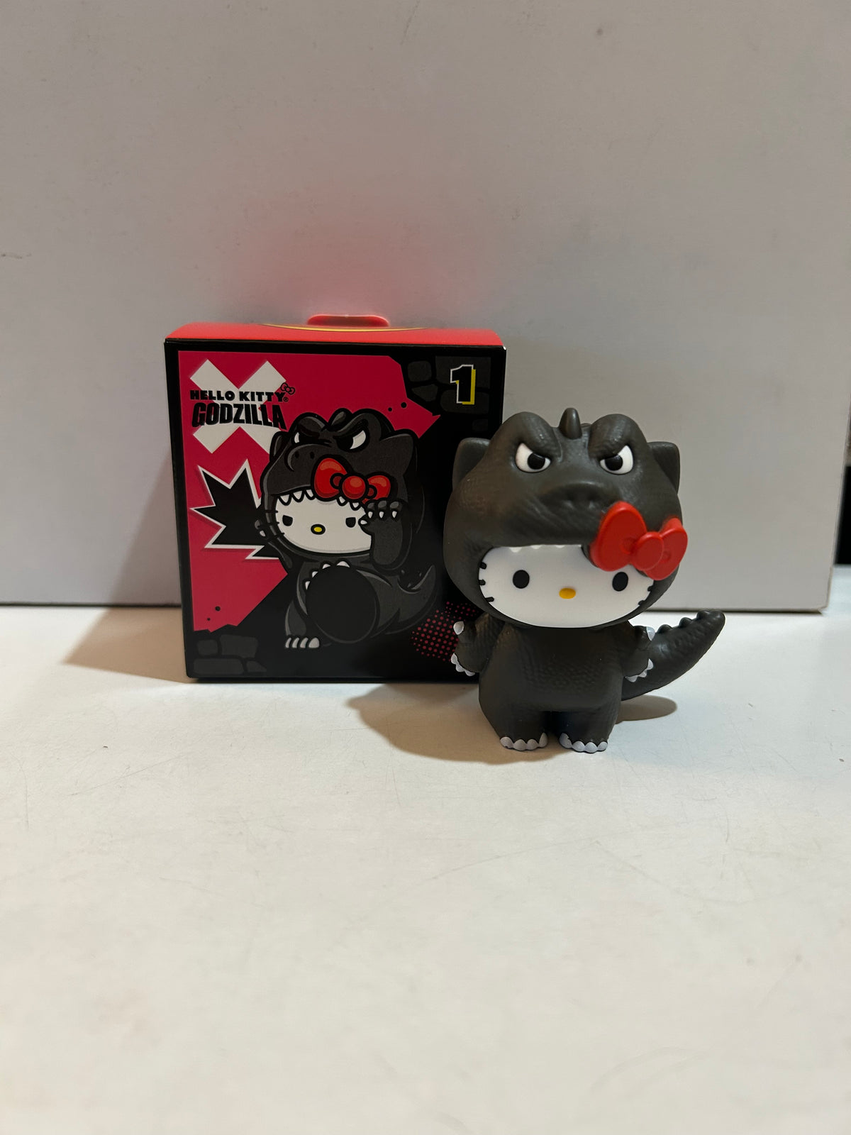 Hello Kitty in a dark Godzilla costume with red bow beside its illustrated box