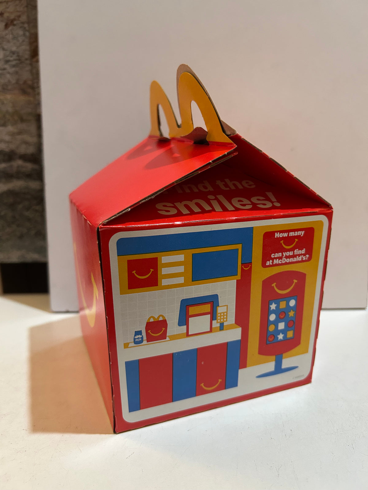 Red McDonald's Happy Meal box with illustrated restaurant counter and kiosk, Find the Smiles activity, and yellow arch handles